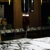 Crescendo Flute Glass filled with a deep purple cocktail garnished with lavender standing on a marble surface and a dark background