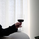 Person holding a glass of red wine against a white curtain background