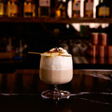 Jazz rum glass with a cocktail with whipped cream and a caramelized banana on a bar counter with blurred bottles in the background