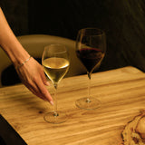 Hand reaching to a Magnifico wine glass filled with white wine next to another Magnifico glass filled with red wine on a wooden table and a dark background