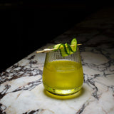 Mixology Cocktail Club DOF glass filled with a bright green cocktail and garnished with a cucumber standing on a marble surface