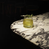 Mixology Cocktail Club DOF glass filled with a bright green cocktail and garnished with a cucumber standing on a marble surface