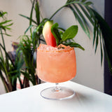 Sublime Cognac glass filled with a light red watermelon cocktail, garnished with mint and a watermelon slice, standing on a white surface and plants in the background