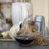 Vinea wine decanter filled with red wine standing on a grey surface and a bouquet of dry flowers in the background