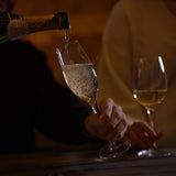Hand holding Vinea Prosecco glass that is being filled with Sparkling wine on a dark background.