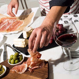Vinoteque Robusto Red Wine Glass filled with red wine standing kitchen table filled with charcuterie and cheeses and a hand organizing a board