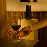 Hand holding Vinoteque Riserva Red Wine glass filled with red wine and standing on a wooden table, wooden wall and a table lamp the background