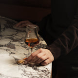 Hand holding Vinoteque Snifter glass filled with brown spirit and standing on a marble surface and a dark background