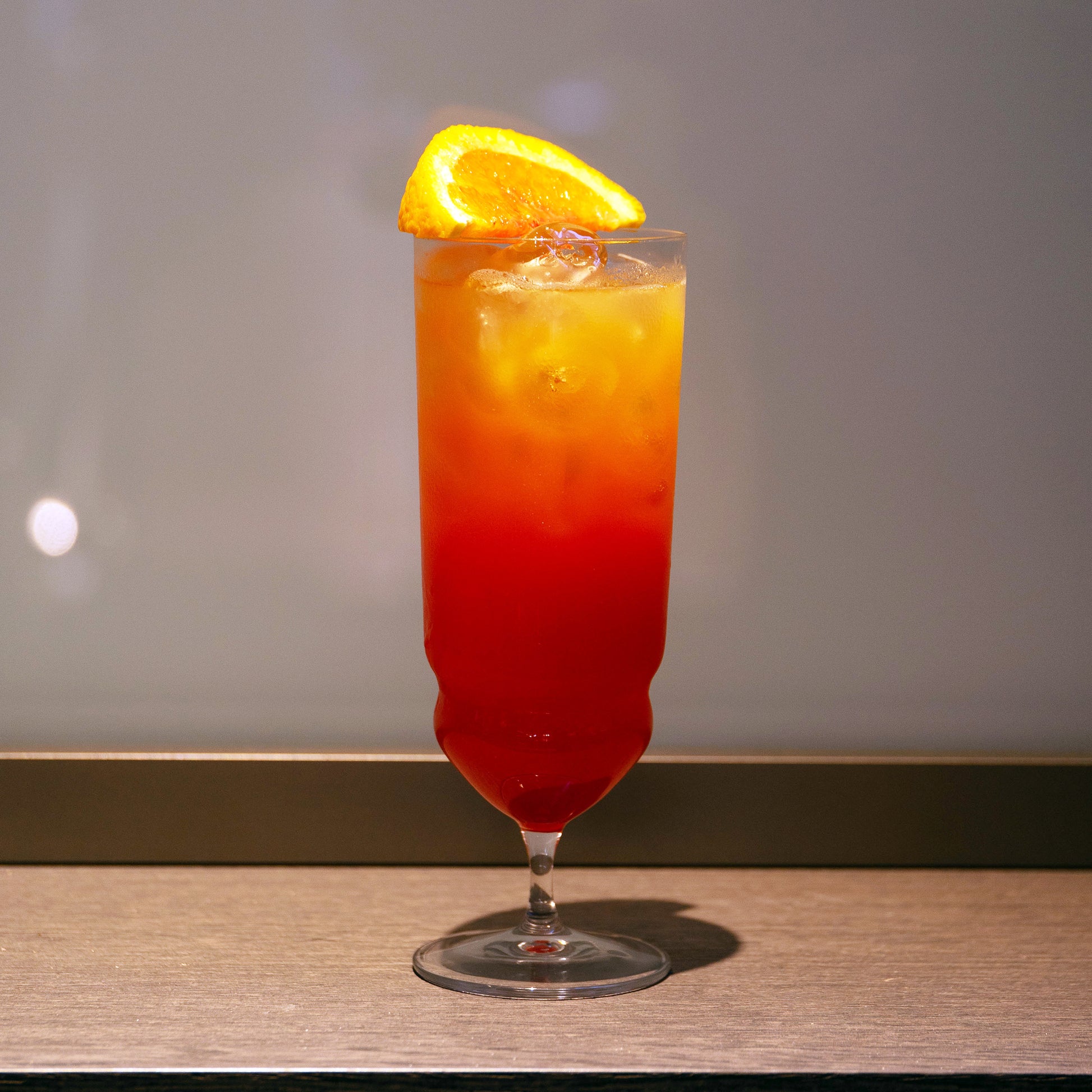 Backdoor '20s Tequila Sunrise Cocktail Glass filled with a bright red cocktail and garnished with a slice of blood orange, standing on a wooden surface and a neutral background
