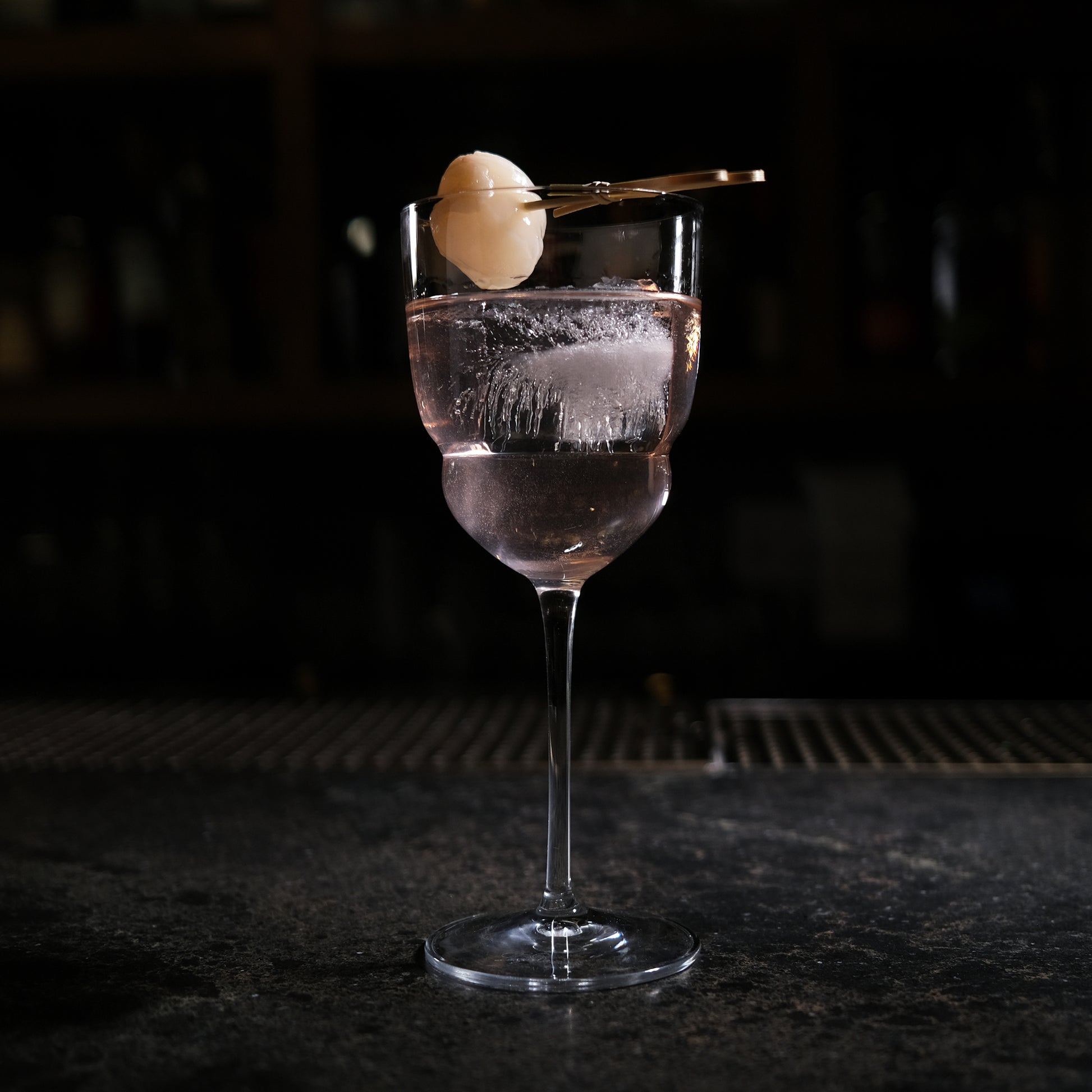 Backdoor '20s Barracuda Cocktail Glass filled with a clear cocktail and garnished with a Lychee on a bar surface and a dark background