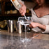 Empty Backdoor '20s Great Gatsby Cocktail Glass standing on a bar surface and a bartender making a cocktail in the background