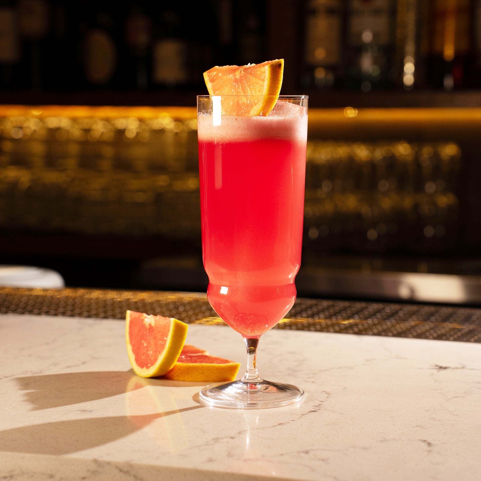 Backdoor '20s Tequila Sunrise Cocktail Glass filled with a bright red cocktail and garnished with a slice of blood orange, standing on a marble surface next to additional blood orange slices and a dark blurred background