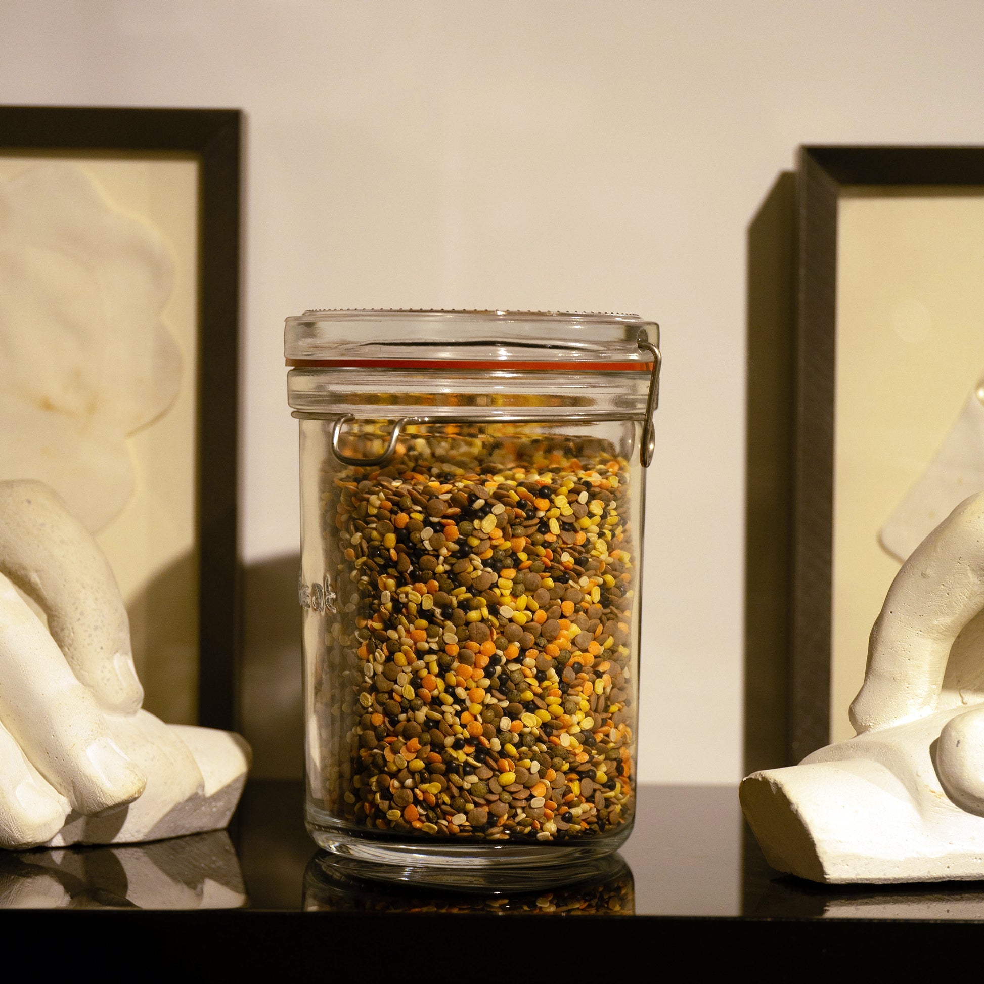 Lock-Eat jar filled with legumes standing on a black reflective surface and frames and hand sculptures in the background