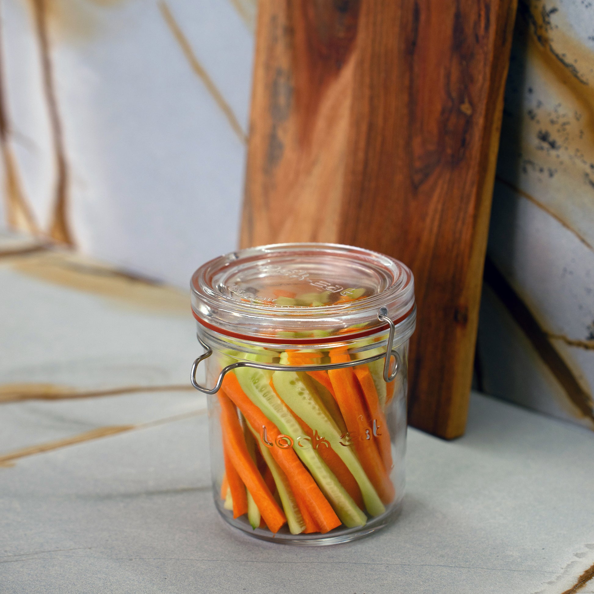 Lock-Eat jar filled with cut cucumbers and carrots standing on a marble surface with a wooden board in the background