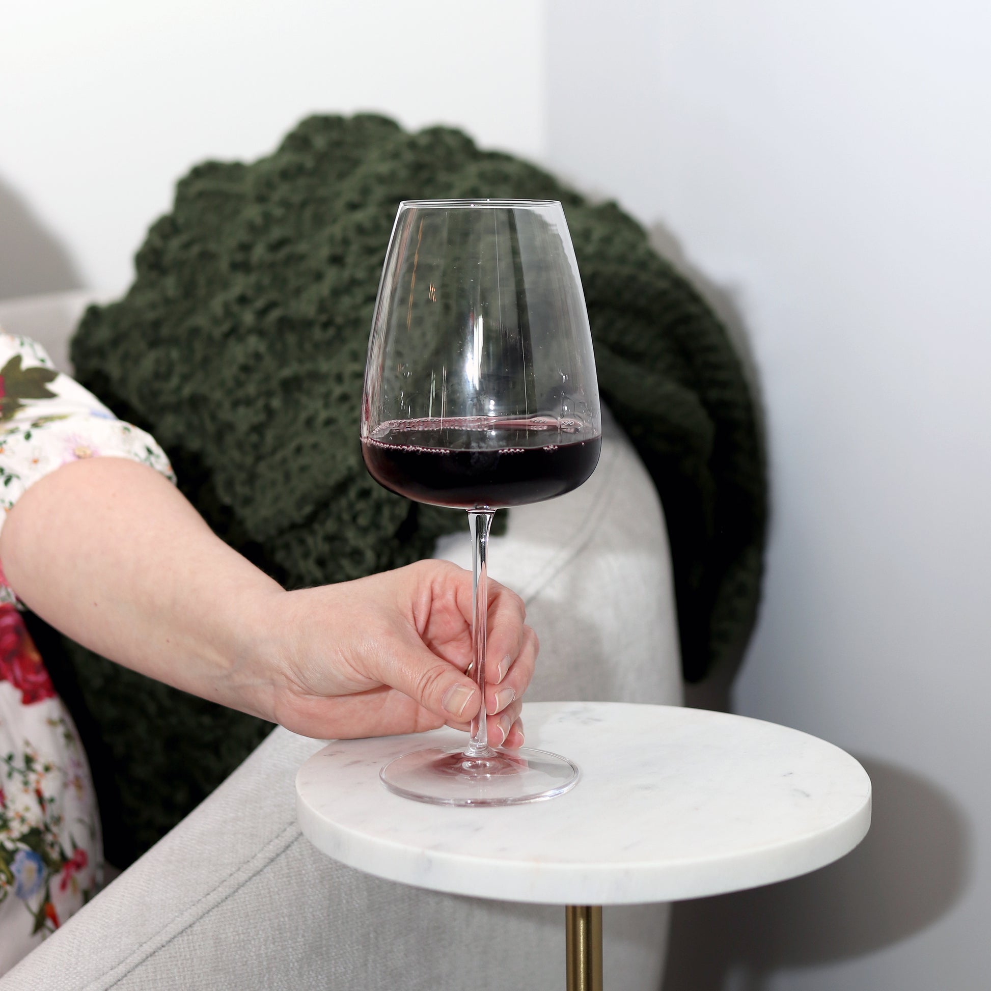 Hand holding a Talismano Italian Premium Bordeaux Red Wine Glass filled with a red wine standing on a white side table with a white chair and a blanket in the background