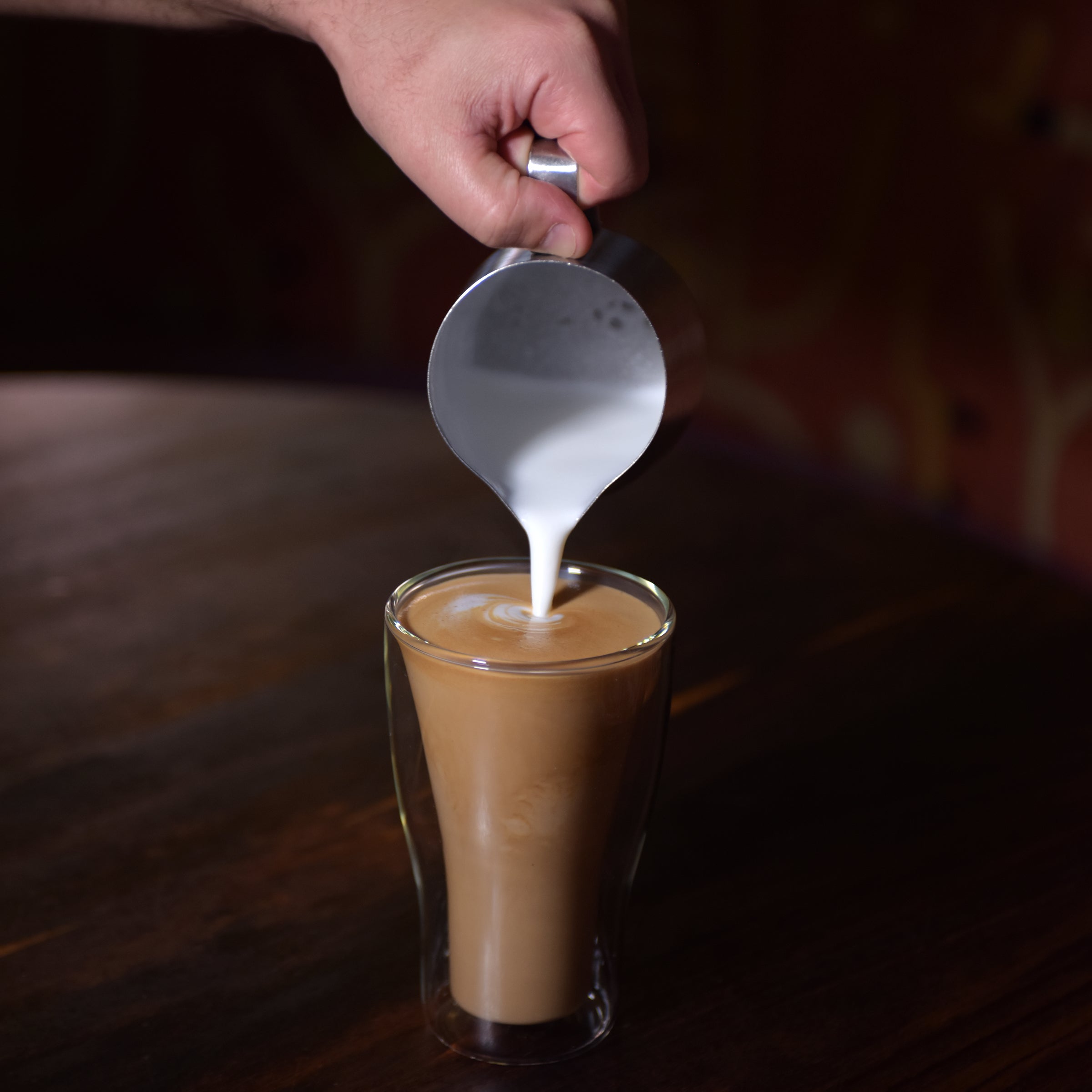 Thermic double-wall latte cup filled with latte and a hand pouring milk inside, standing on a dark wooden surface