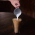 Thermic double-wall latte cup filled with latte and a hand pouring milk inside, standing on a dark wooden surface