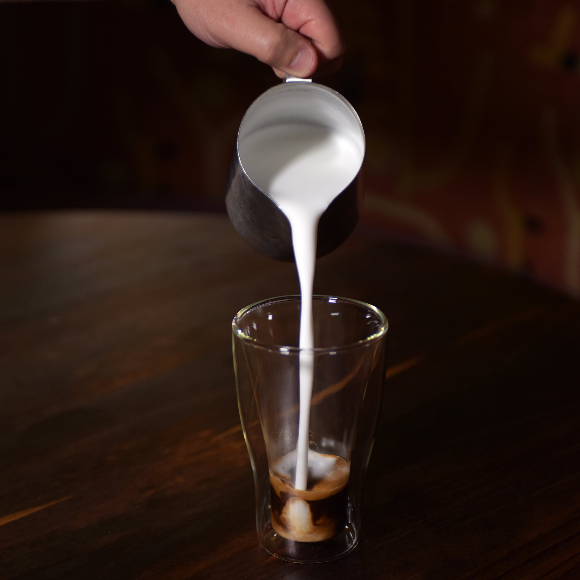 Thermic double-wall latte cup filled with latte and a hand pouring milk inside, standing on a dark wooden surface
