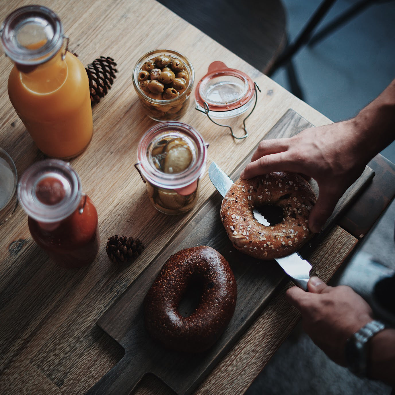 Filled Lock-Eat jars with juice and food on a wooden kitchen table, while two hands with a knife, cutting bagels on a wooden board. 