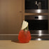 Atelier stemless glass filled with bright red cocktail and garnished with apple and thyme standing on a wooden surface and blurred kitchen appliances in the background