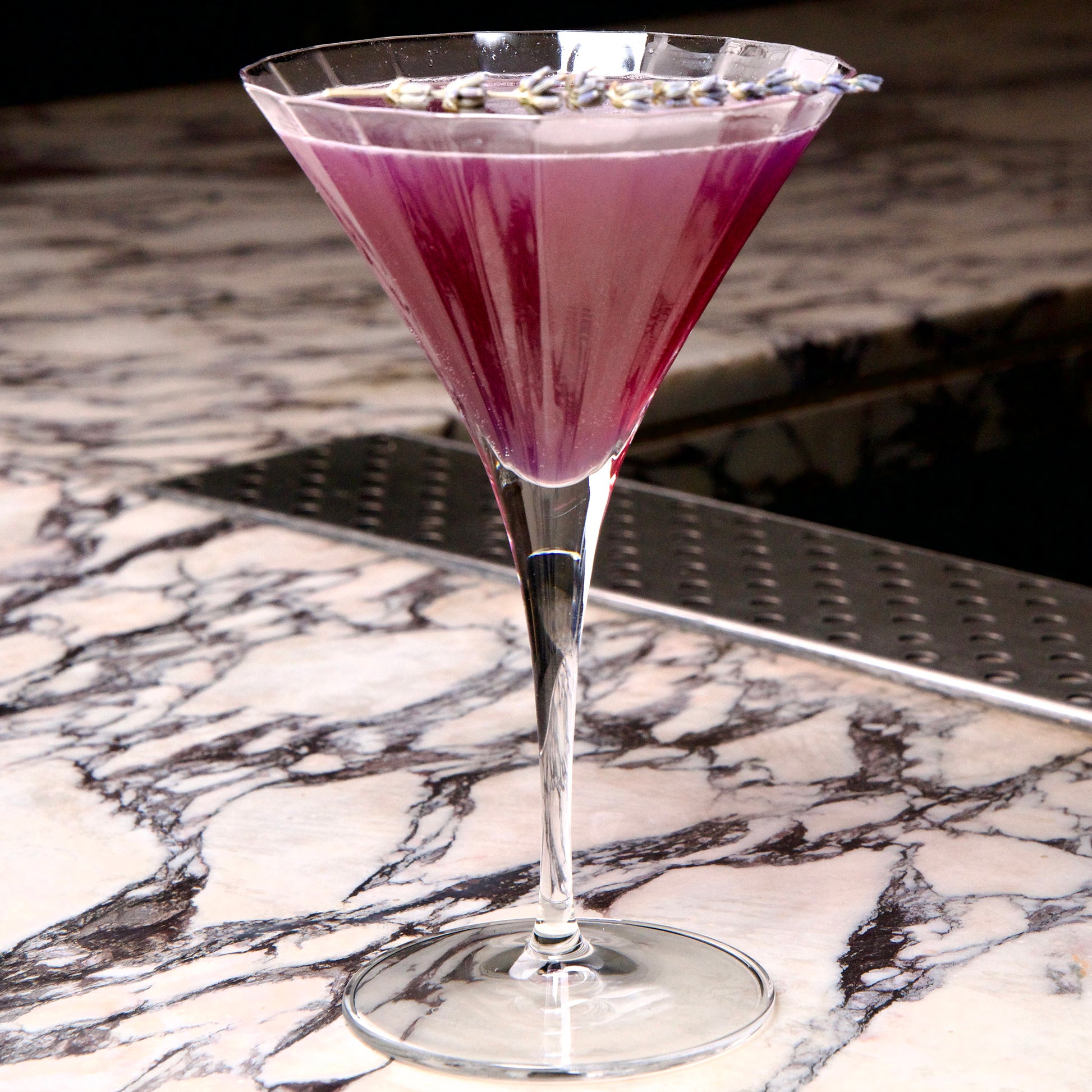 Bach Martini glass with a deep purple cocktail garnished with lavender standing on a black and white marble surface