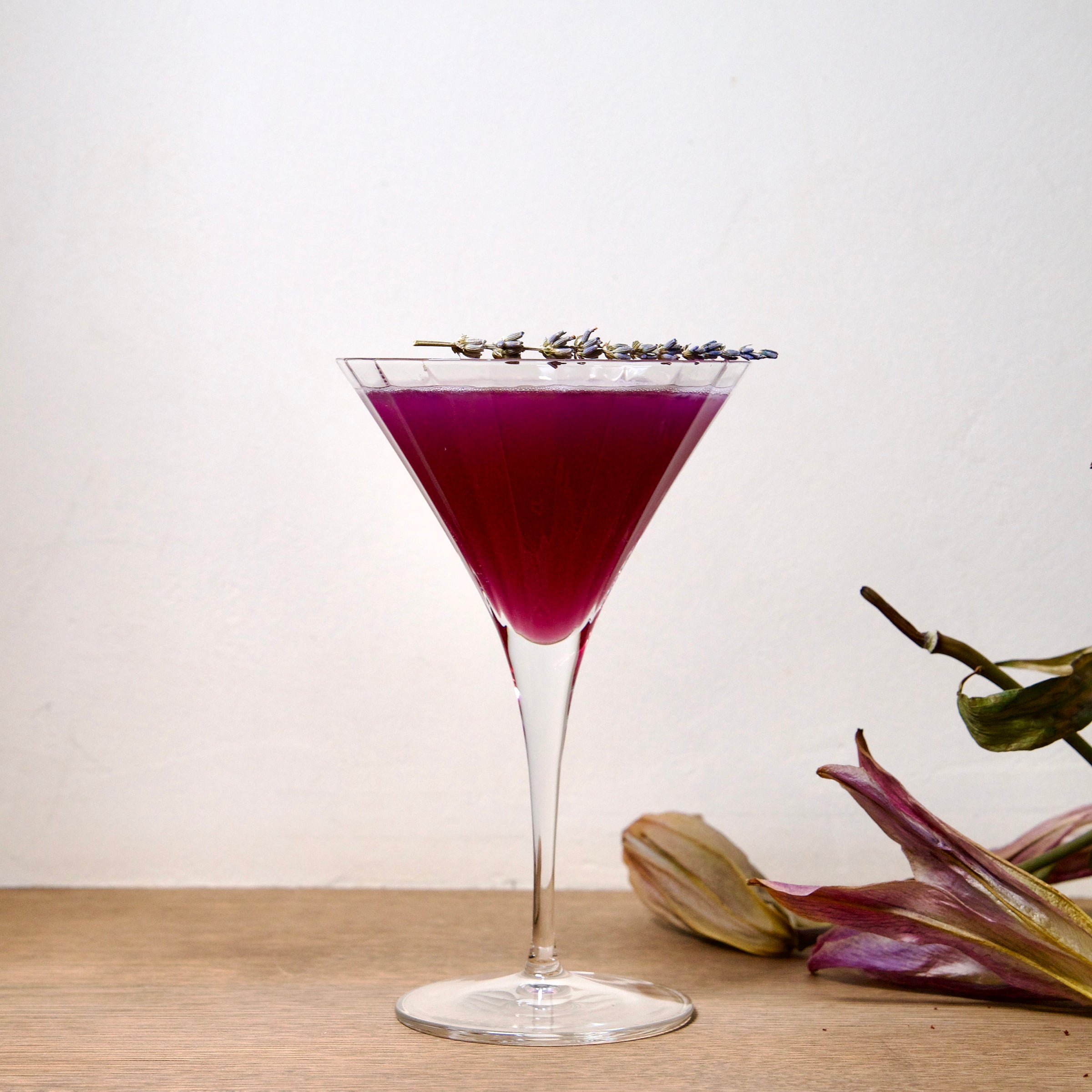 Bach Martini glass with a deep purple cocktail garnished with lavender standing on a wooden surface next to flowers and a white wall in the background