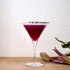 Bach Martini glass with a deep purple cocktail garnished with lavender standing on a wooden surface next to flowers and a white wall in the background