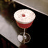 Bach Retro Fizz Cocktail glass with a red cocktail and white foam garnished with a raspberry standing on a dark surface