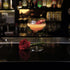Backdoor '20s Great Gatsby glass with a orange cocktail garnished with a single red rose petal on a dark bar surface next to a red rose with blurred bottles in the background