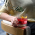 Person holding a glass of non-alcoholic cocktail with a lemon twist and olive branch, sitting on a wooden chair.