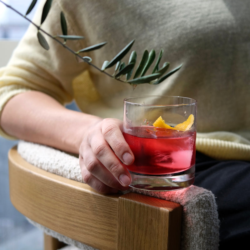 Person holding a glass of non-alcoholic cocktail with a lemon twist and olive branch, sitting on a wooden chair.