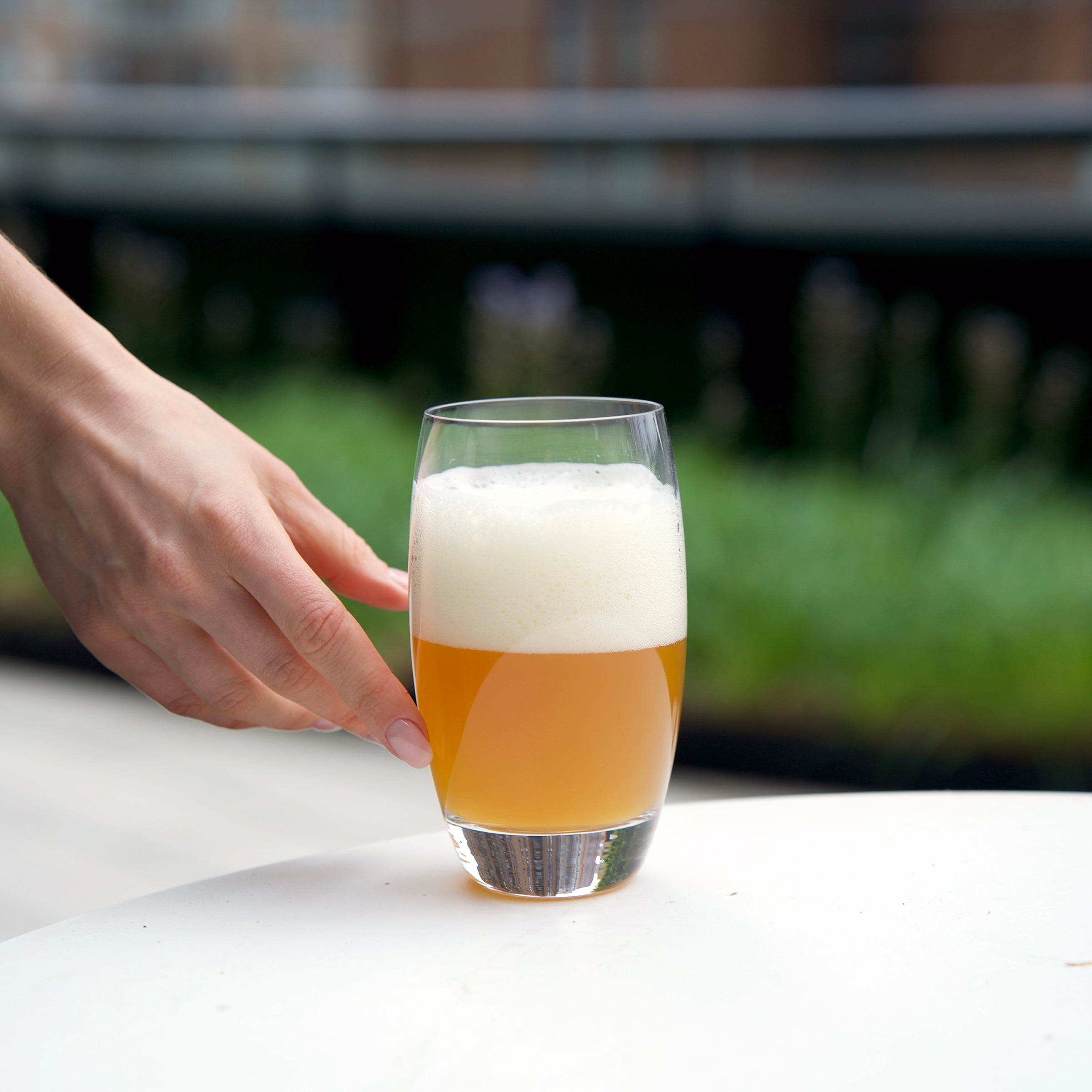 hand reaching a Crescendo Highball drinking glass half filled with beer and thick foam, standing on a white surface and blurred background