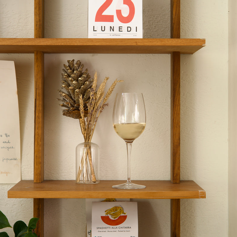 Wooden shelf with a glass of white wine, decorative vase, and calendar on a neutral wall.