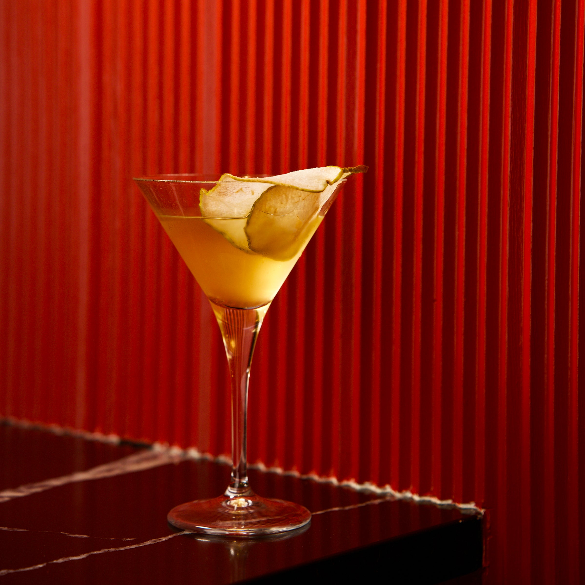 Cocktail garnished with a pear, in a Crescendo martini glass with a red wall background
