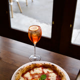 Magnigico Small glass with Spritz cocktail with a slice of orange next to a pizza on a wooden table.