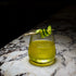 Mixology Cocktail Club DOF glass filled with a bright green cocktail and garnished with a cucumber standing on a marble surface