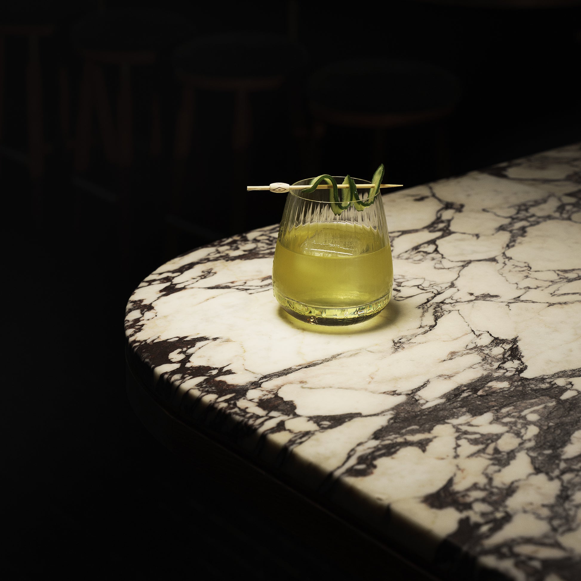 Mixology Cocktail Club DOF glass filled with a bright green cocktail and garnished with a cucumber standing on a marble surface