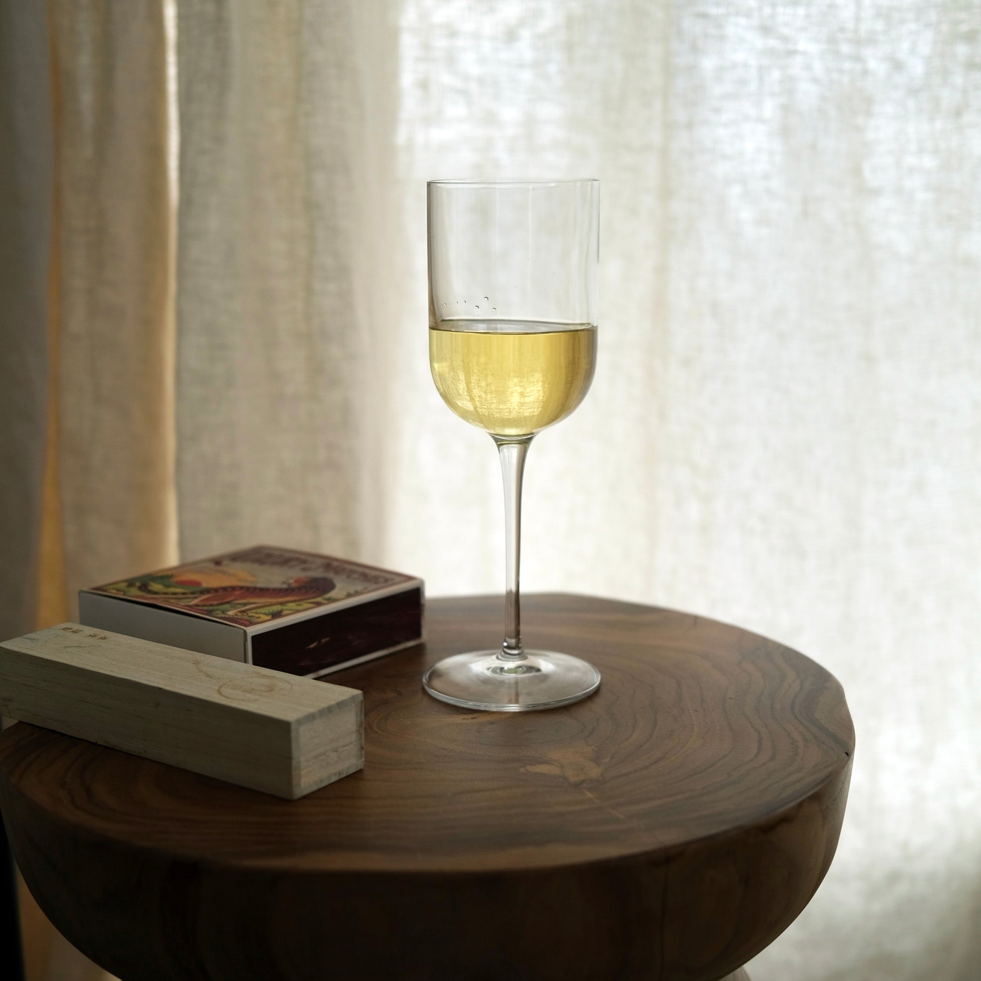 Glass of white wine on a wooden table with a book and decorative box against a curtain background