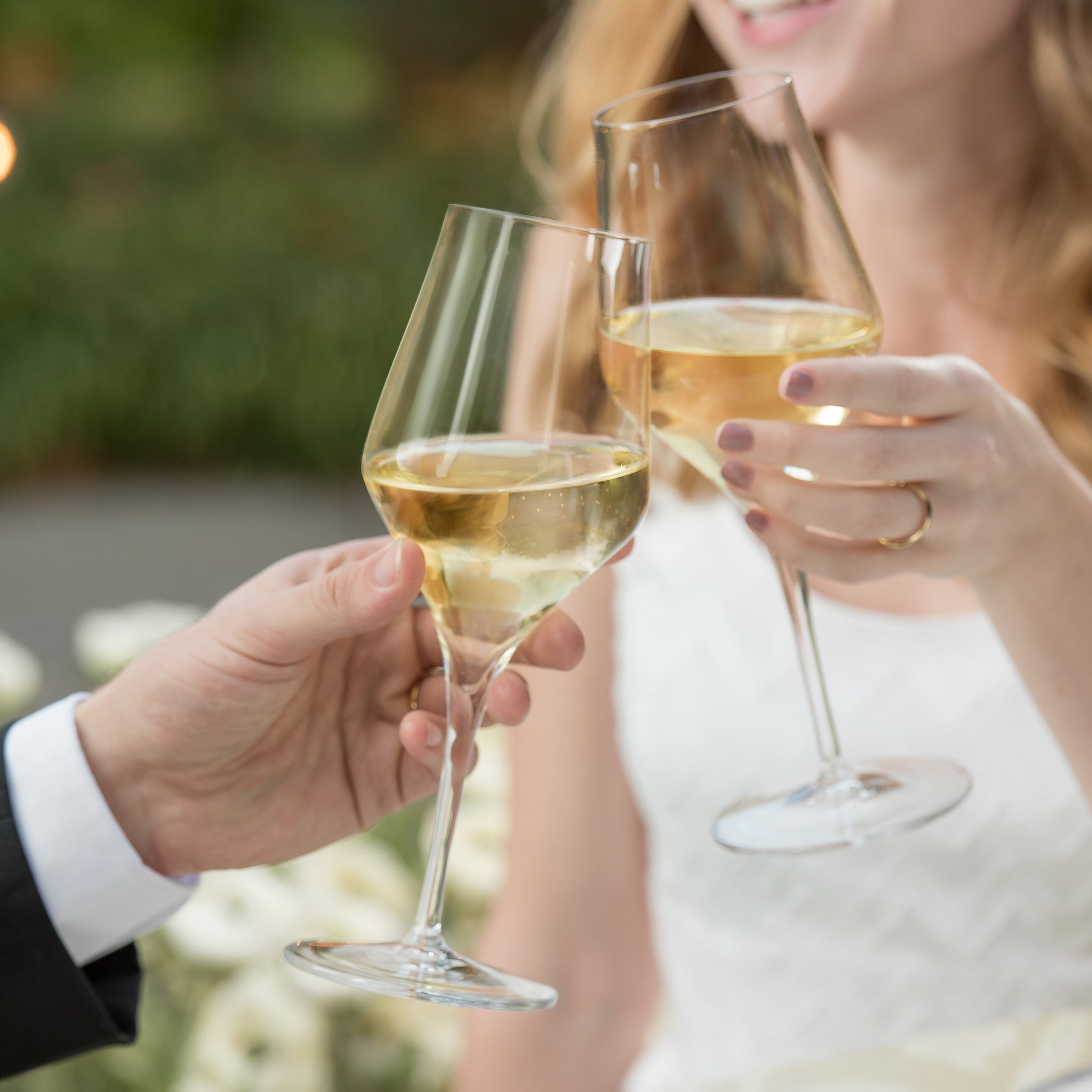 Two people clinking Supremo Chardonnay wine glasses filled with sparkling wine with a blurred outdoor background