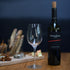 Empty Vinoteque Red Wine Glass standing next to a red wine bottle on a wooden table, behind is a blurred cheese board and a dark background