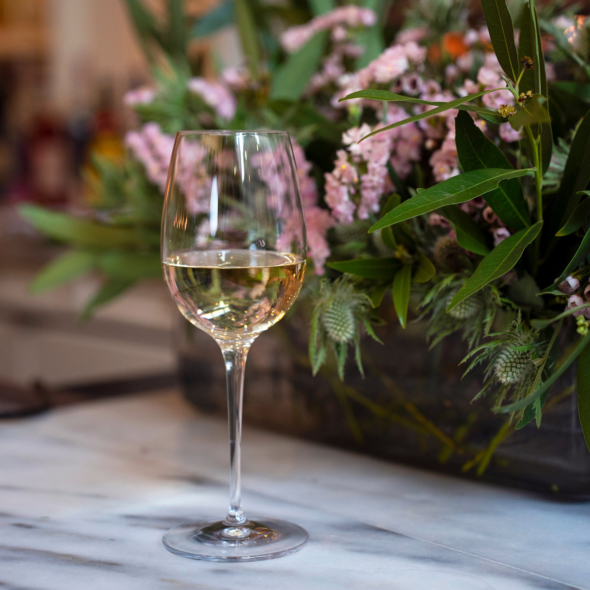 Vinoteque Red Wine Glass filled with white wine standing on a marble surface and a bouquet of pink flowers and greenery in the background