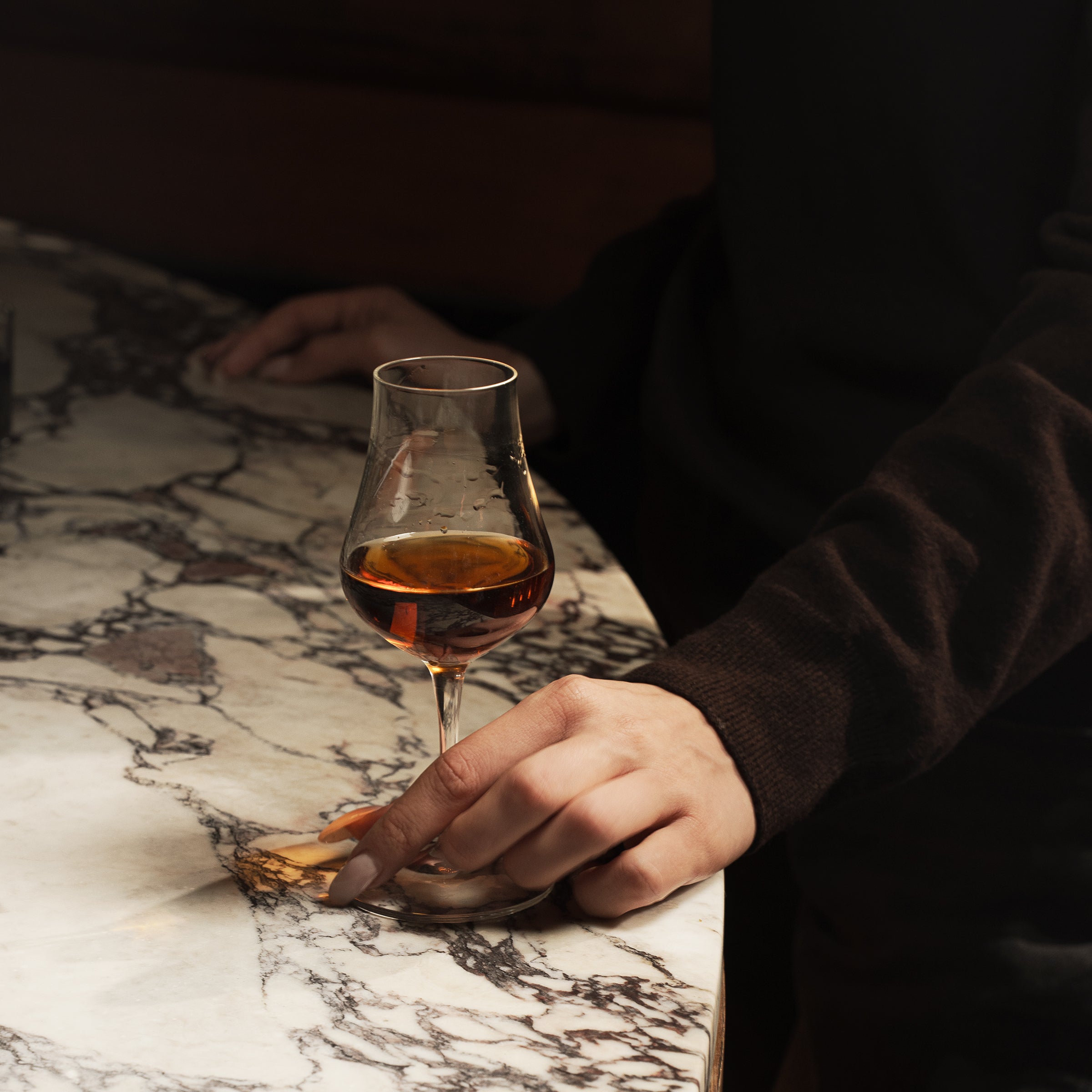 Hand holding Vinoteque Snifter glass filled with brown spirit and standing on a marble surface and a dark background